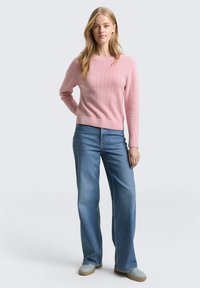 Różowy sweter dzianinowy z teksturowanym wzorem, okrągłym dekoltem i prążkowanym ściągaczem. Połączony z jasnoniebieskimi, szerokimi spodniami dżinsowymi i szarymi butami wsuwanymi.