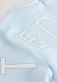 Heller blauer, strukturierter Stoff mit großen, erhabenen weißen und silbernen Buchstaben. Reißverschluss hinten. Glattes, modernes Design.