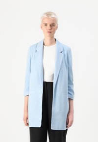 Ανοιχτό μπλε σακάκι με υφή, μονόπετο σχέδιο και δύο μπροστινές τσέπες, φορεμένο πάνω από άσπρο πουκάμισο και μαύρο παντελόνι.