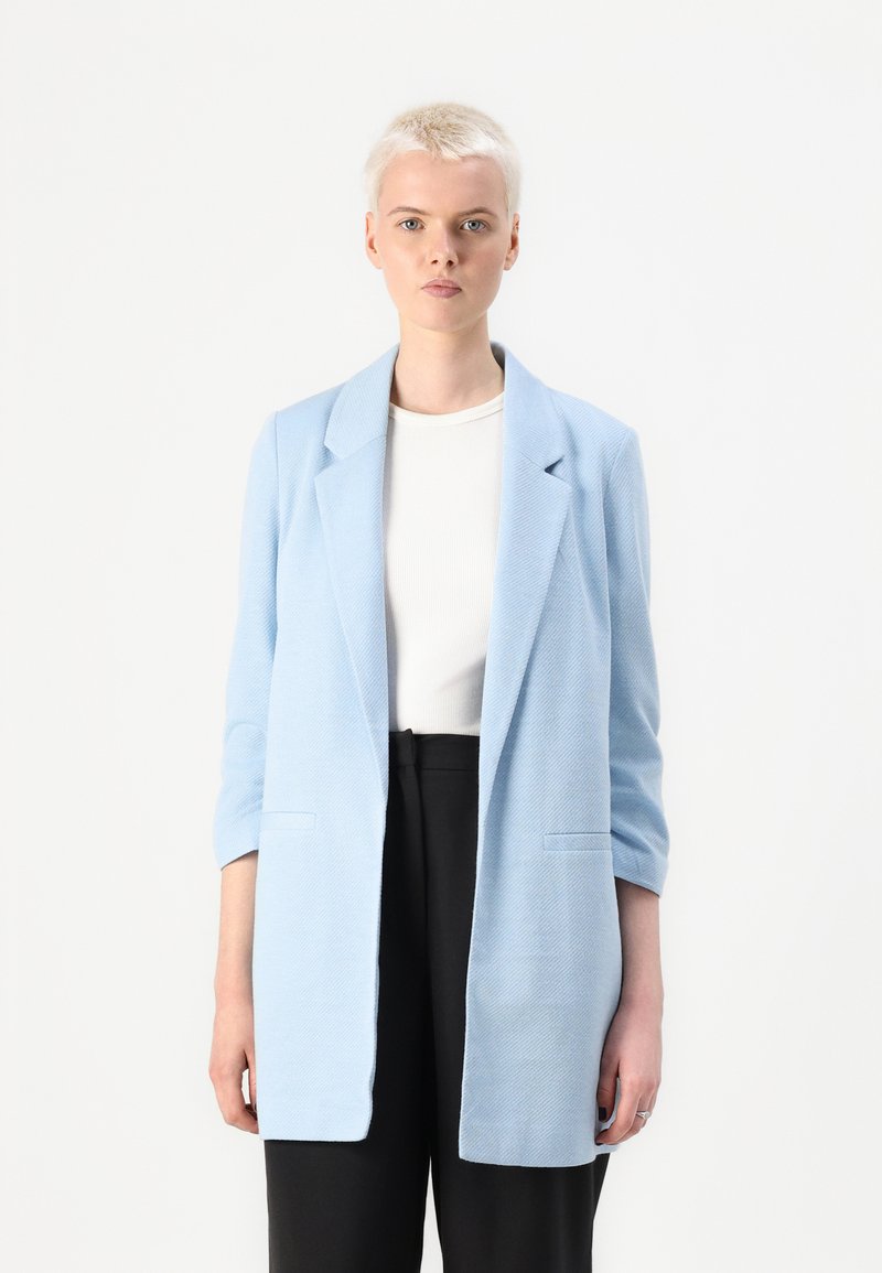 Ανοιχτό μπλε σακάκι με υφή, μονόπετο σχέδιο και δύο μπροστινές τσέπες, φορεμένο πάνω από άσπρο πουκάμισο και μαύρο παντελόνι.