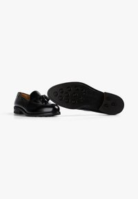 Mocasines de cuero negro con un acento de borlas, punta redondeada y suela de goma texturizada con patrones circulares. Acabado elegante y pulido.
