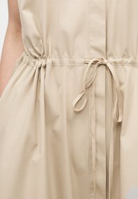 Κοντινό πλάνο ενός μπεζ φορέματος με σφικτή μέση που δένει με λεπτό υφασμάτινο κορδόνι σε φιόγκο.