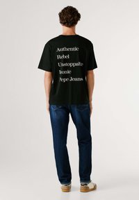 Camiseta negra con un gran texto blanco en la parte trasera: "Auténtico, Rebelde, Imparable, Icónico, Pepe Jeans." Combínala con jeans de mezclilla azul.