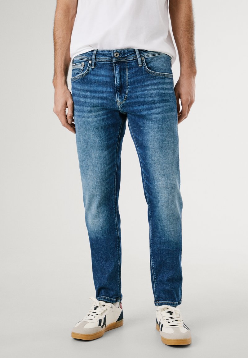 Blue denim jeans with a slim fit, featuring subtle fading, five pockets, and a zip fly. Paired with white sneakers.