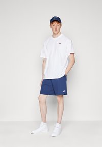 Maglietta bianca, pantaloni corti blu navy e sneaker bianche. La maglietta presenta un piccolo logo rosso; i pantaloni corti hanno un logo bianco all'orlo. Il cappellino abbina i pantaloni corti.