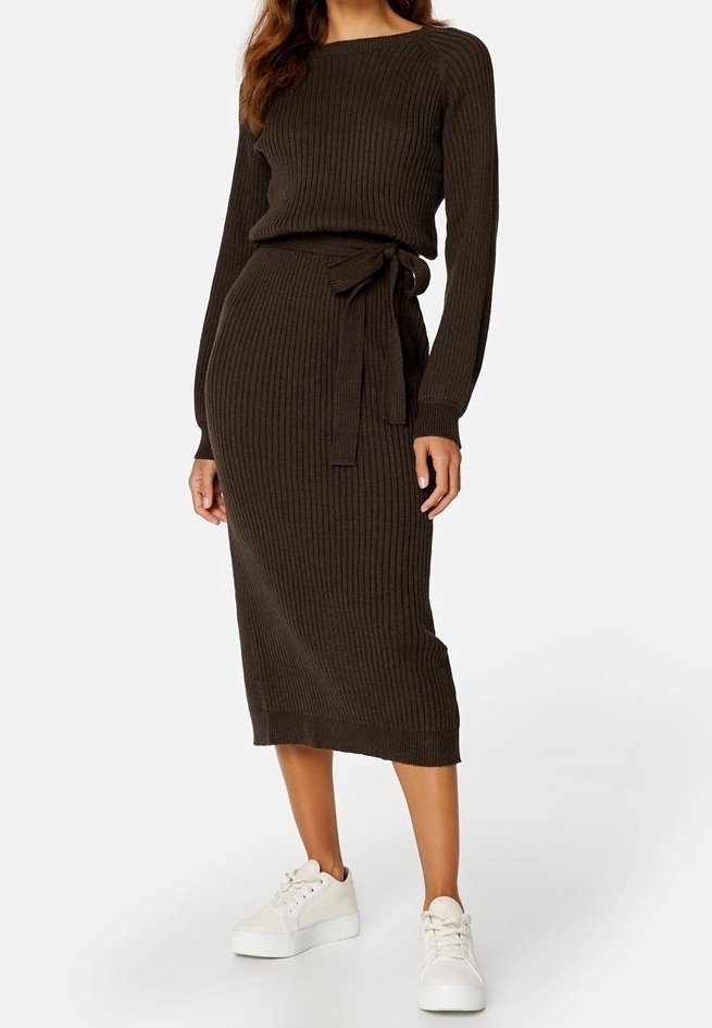 Femme portant une robe marron foncé en maille côtelée à manches longues avec une ceinture à la taille, associée à des baskets blanches à plateforme, debout devant un fond blanc.