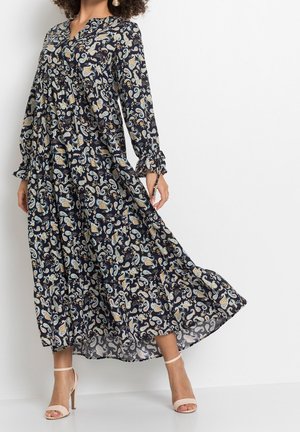 Kobieta ubrana w długą granatową sukienkę w wzór paisley z długimi rękawami i beżowe sandały na wysokim obcasie, stojąca na tle jednolitego białego tła.