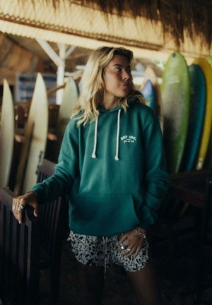 Bluza z kapturem w kolorze zielonkawoniebieskim (teal) z białymi troczkami i logo, zestawiona z czarno-białymi szortami we wzory. W tle widoczne deski surfingowe. Obok drewniane meble.