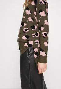 Vista laterale di una persona che indossa un maglione verde oliva con macchie rosa e nere e pantaloni in pelle neri, su uno sfondo neutro.