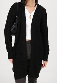 Mustat neulottu neulecardigan, jossa on palmikkokuvio, avoin etuosa ja pitkät hihat. Päällä valkoinen paita, yhdistettynä tummaan housupariin.