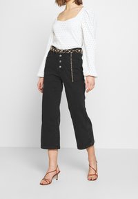 Vaqueros negros cortos con cuatro botones delanteros, combinados con una blusa blanca de ojales y un cinturón de cadena dorada. Sandalias rojas de tiras completan el conjunto.