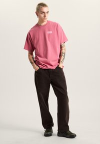 Lyserød bomulds T-shirt med et lille hvidt logo. Kombinationen med mørkebrune bukser og sorte og grønne sneakers. Modellen har kort hår og tatoveringer.