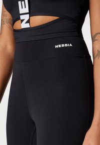 Black high-waisted leggings with NEBBIA logo. Smooth, stretchy fabric. Features cut-out design and seams for a fitted look.