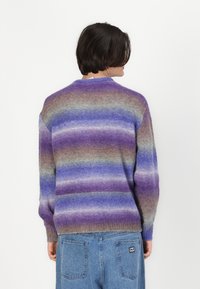 Persoon met kort donker haar, gekleed in een fuzzy paarse, blauwe en bruine gestreepte trui en blauwe denim broek, met de rug naar de camera op een witte achtergrond.