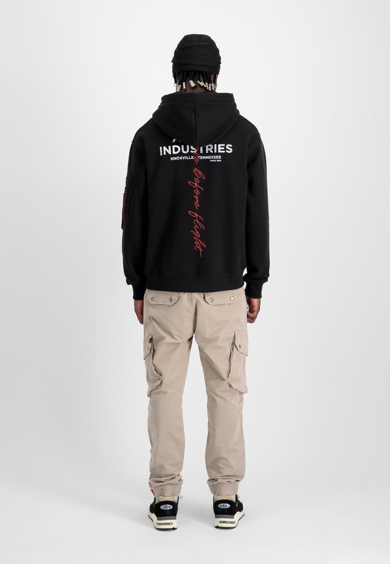 Schwarzer Kapuzenpullover mit weißem und rotem Text auf der Rückseite, kombiniert mit beigen Cargo-Hosen mit Seitentaschen und schwarzen Turnschuhen.