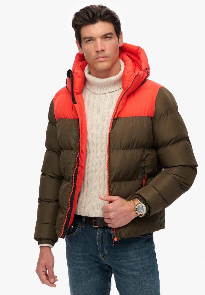 Padded Jacke in olivgrün und leuchtend orange, mit Kapuze, Reißverschlusstaschen und Kontrastnähten. Getragen über einem cremefarbenen Pullover.