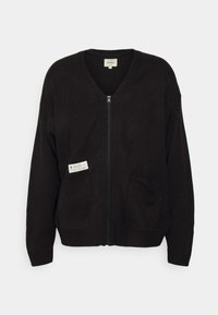 Cárdigan negro de punto con escote en V, cremallera frontal, dos bolsillos y una pequeña etiqueta en el bolsillo izquierdo. Puños y dobladillo acanalados.