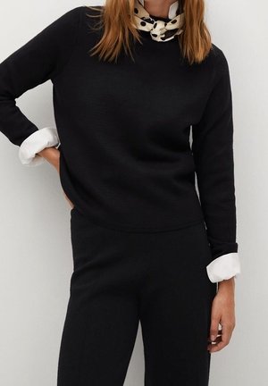 Kvinde iført sort langærmet sweater og bukser, hvide skjortefmanchetter synlige, med et plettet halstørklæde.