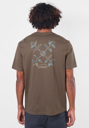 Personne aux cheveux bouclés portant un t-shirt marron avec un motif de croix florale bleu et jaune avec le mot "OXBOW" dans le dos.