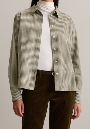 Femme portant une chemise à carreaux olive boutonnée sur un col roulé blanc rentrée dans un pantalon en velours côtelé marron avec une ceinture noire.