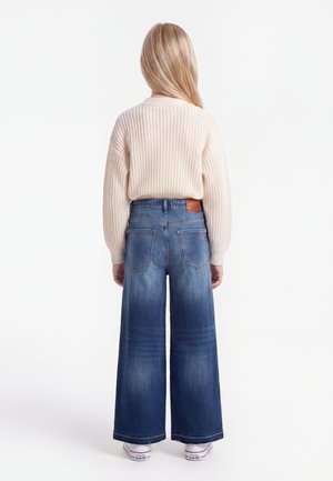 Barn med langt blondt hår står med ryggen til iført en cremefarvet strikket sweater, vidde blå jeans og hvide sneakers.