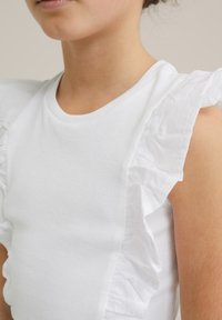Bela rebrasta majica na bretele s nabranim belim tekstilnim detaljima na ramenima. Tekstura kontrastira s gladkim tijelom majice.