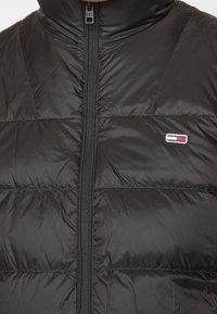 Czarna puchowa kurtka z wysokim kołnierzem, poziomym pikowanym wzorem i zamkiem z przodu. Posiada małe logo na wysokości klatki piersiowej.