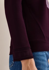 Felpa borgogna con polsini a coste, texture liscia e dettaglio sulla cucitura laterale. Abbinata a jeans in denim blu.