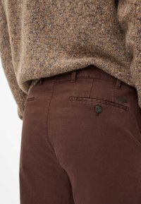 Braune Chinos mit glatter Textur, ausgestattet mit einer einzelnen Knopftasche und Gürtelschlaufen, kombiniert mit einem strukturierten braunen Strickpullover.