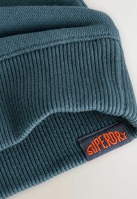 Berretto a coste in teal con risvolto e un'etichetta nera in tessuto con il testo arancione "SUPERDRY" ricamato sul bordo.