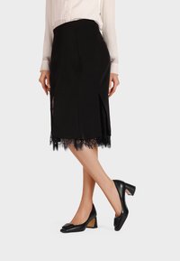 Gonna nera a matita con rifiniture in pizzo all'orlo, caratterizzata da spacchi laterali. Abbinata a scarpe nere con tacco blocco, punta quadrata e dettaglio a fiocco.