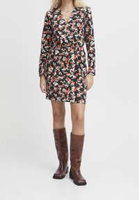 Robe à imprimé floral noire avec des accents rouges, blancs et verts ; décolleté en V, manches longues et taille élastique. Portée avec des bottes en cuir marron jusqu'au genou.