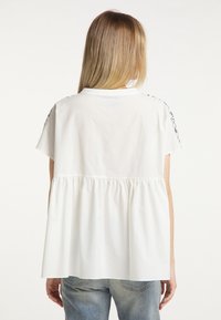 Chemise en coton blanc avec une coupe décontractée, un dos froncé, des manches courtes et des surpiqûres noires décoratives le long des coutures d'épaules.