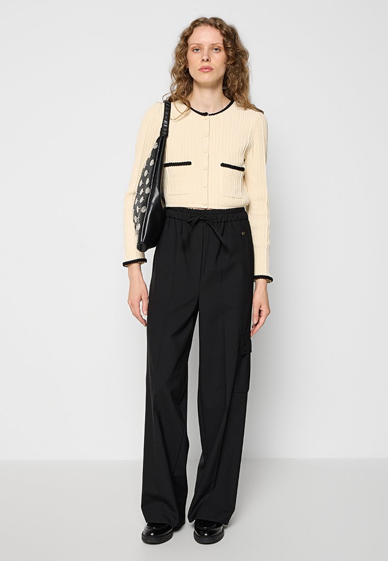 Beige geribde cardigan met zwarte randen en zakken, gecombineerd met losvallende zwarte broek. Model houdt een zwarte tas met ronde accenten vast.