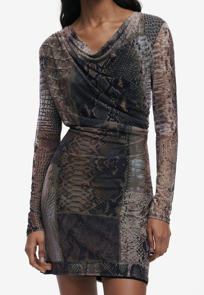 Femme portant une mini-robe moulante à manches longues avec des motifs en peau de serpent marron et noir mélangés et un décolleté drapé.