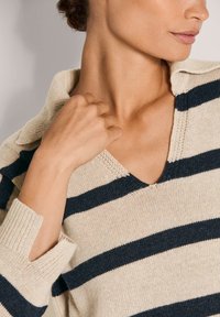 Frau trägt einen beigen und schwarzen gestreiften V-Ausschnitt-Pullover mit Kragen, hält den Pullover am Halsbereich und schaut nach rechts.