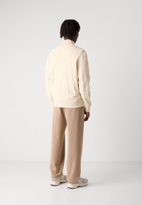 Man som står med ryggen mot kameran iförd en krämfärgad texturerad tröja, beige vida byxor och vita sneakers mot en enfärgad bakgrund.