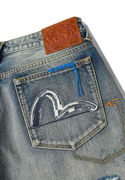 Tasca posteriore di jeans in denim con design astratto ricamato in bianco e blu navy, dettaglio in filo blu e etichetta del marchio in pelle marrone.