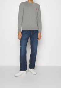 Szary sweter dziany z okrągłym dekoltem i czerwonym logo, zestawiony z ciemnoniebieskimi jeansami i białymi tenisówkami. Teksturowana tkanina i klasyczny krój.