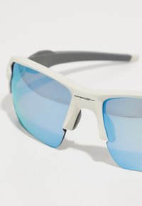 Weiße und graue Sportsonnenbrille mit blauen Verlaufsobjektiven, kantigem Rahmen-Design und gummierter Textur an den Nasenpads und Bügeln.
