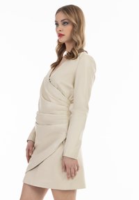 Rochie din piele bej cu mâneci lungi, decolteu în V și design fiorit. Prezintă un finisaj texturat și o linie de tiv asimetrică, suprapusă.