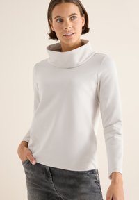 Langärmliges, weißes Oberteil mit hohem, umgeschlagenem Kragen. Weicher Stoff mit glatter Textur, kombiniert mit grauen Jeans. Schlichtes und modernes Design.