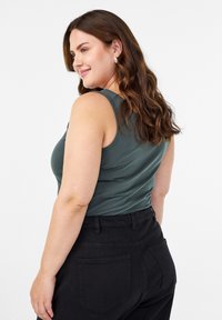 Zelena majica bez rukava s okruglim izrezom, s glatkom teksturom. Uparena s crnim hlačama viskog struka s stražnjim džepovima.