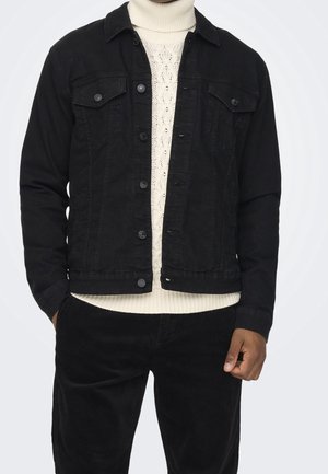 Chaqueta vaquera negra sobre suéter de cuello alto de punto crema, combinada con pantalones negros, manos en los bolsillos, de pie contra un fondo sencillo.