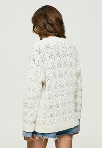 Bílý pletený cardigan s krajkovým vzorem, žebrovanými manžetami a lemem a volným střihem, nošený přes džínové šortky. Pohled zezadu.