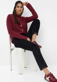 Burgunderfarbener Cardigan mit Holzknöpfen, kombiniert mit schwarzen Cordhosen und kastanienbraunen Sneakern, sitzt auf einem weißen Stuhl.