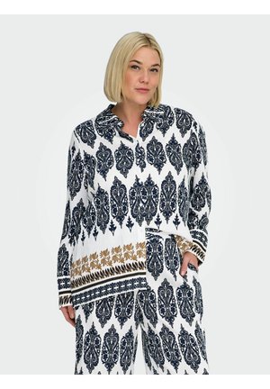 Blondynka ubrana w białą koszulę z długim rękawem oraz spodnie z czarno-złotymi, misternymi, symetrycznymi wzorami, stojąca z jedną ręką w kieszeni.