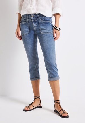 Vrouw draagt lichtblauwe opgerolde cropped jeans, witte blouse met lange mouwen, zwarte platte sandalen met bandjes en een zwart-witte armband aan de rechterpols.