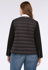 Kvinde med brunt hår, der vender væk, iført en sort quiltet jakke med lynlåse på ærmerne og blå jeans foran en ensfarvet baggrund.