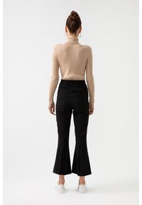 Světle béžový žebrovaný rolák v kombinaci s černými kalhotami s vysokým pasem a rozšířenými nohavicemi, které mají rozparky na lemu. Viditelné bílé boty.